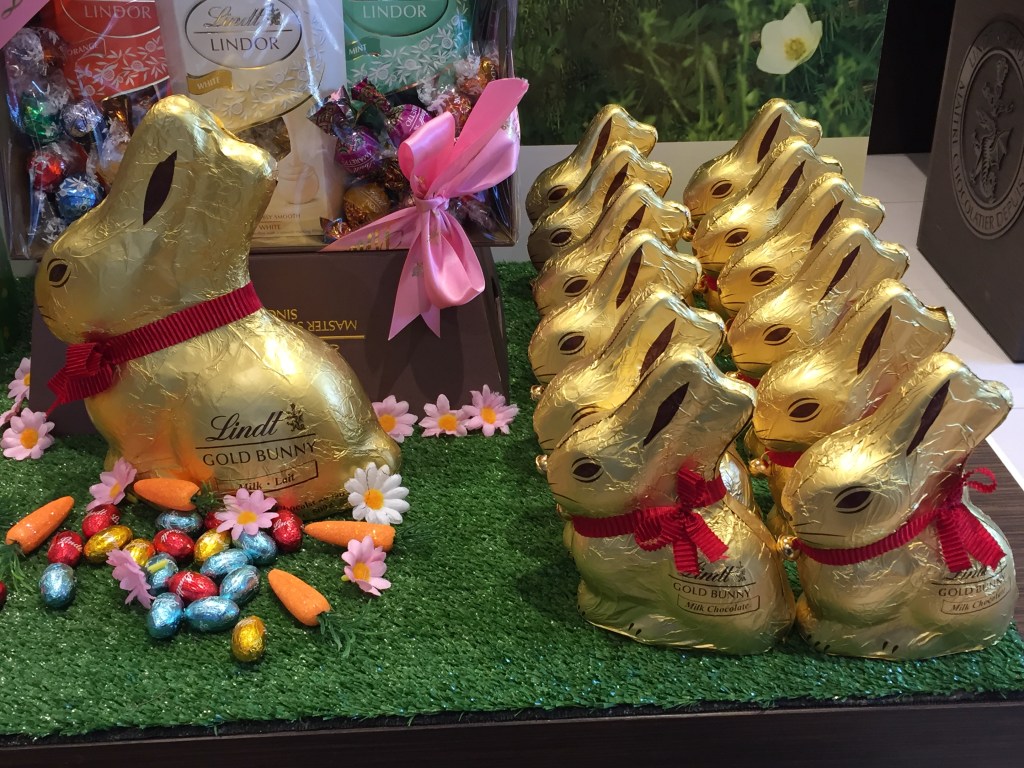 Thirteen Lindt Gold Bunnies - milk chocolate rabbits wrapped in decorative gold foil, sporting red collars with bells - sitting on a patch of fake grass as part of a shop display. Twelve bunnies are regular-sized; the thirteenth is HUGE.