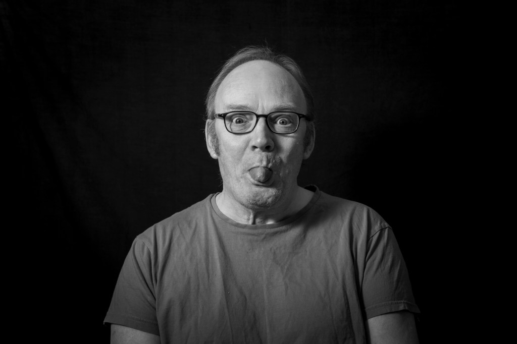 A black-and-white photograph of Simon, a white man with glasses, sticking his tongue out at the camera.