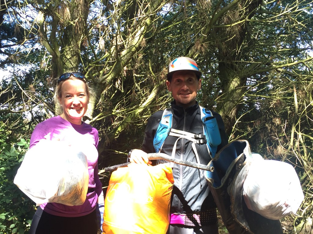 A man and a woman in running clothes stand in a forest, holding up three full plastic bags and other litter.