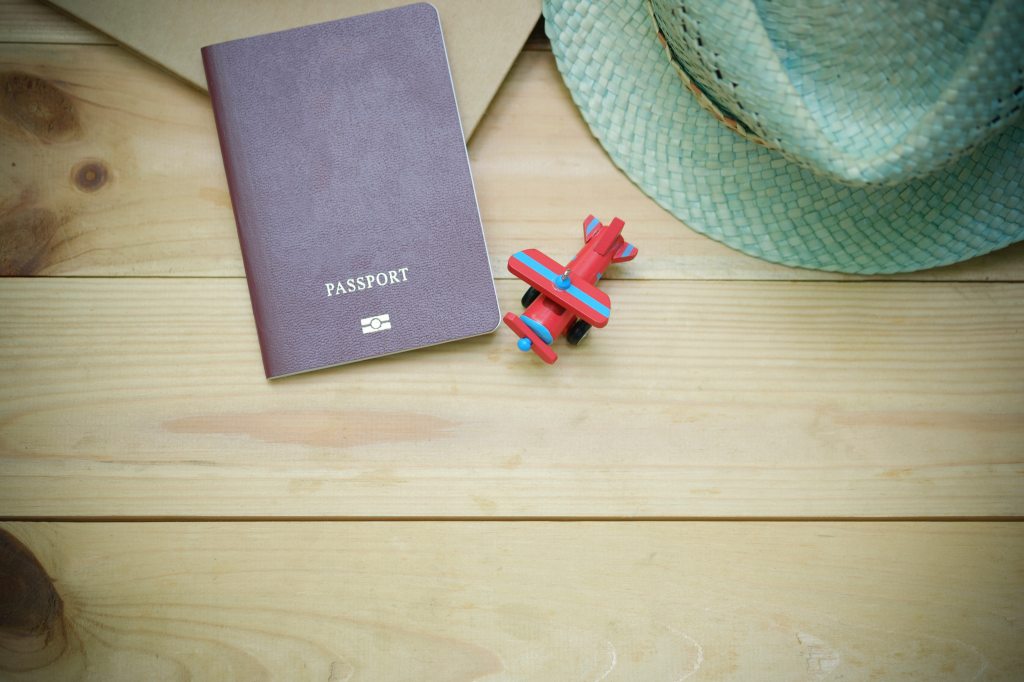 A countryless passport lies on a wooden table next small toy plane and a straw hat.