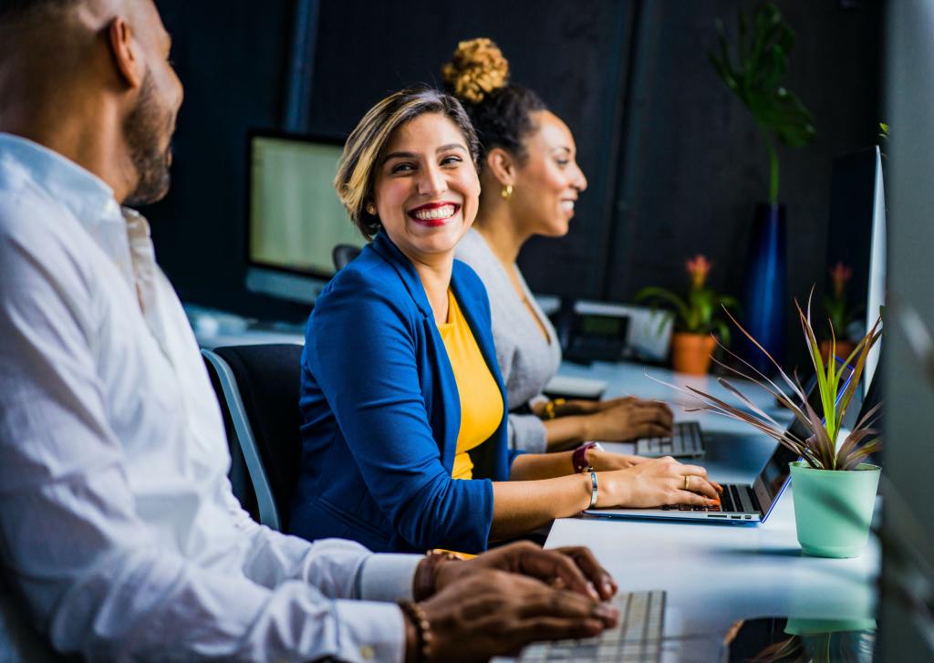 Three office workers laugh with each other while working on laptops