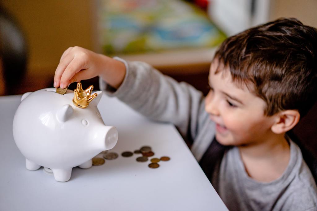 A young boy puts coins into a piggy bank, which is wearing a small golden crown.