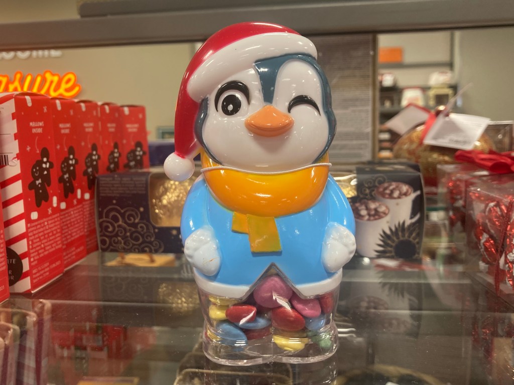 A hollow plastic penguin that holds Smarties-like sugar-coated chocolates. The penguin is wearing a Santa hat and winking at the camera